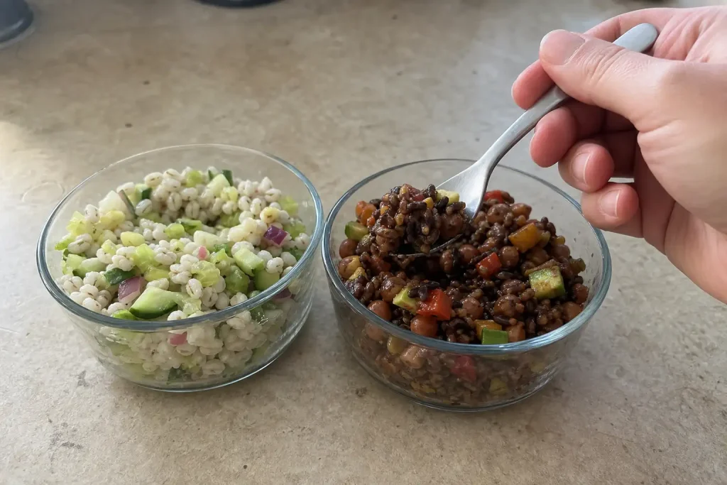 En hand tar upp mat med en sked ur ett av två skålar med olika grönsaks- och spannmålsblandningar på ett köksbord.